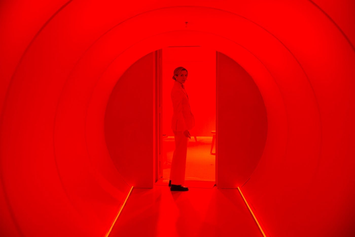 A woman stands at the end of a tube shaped hallway in profile, her head turned towards us. The hallway is painted white and the woman wears white, save for black shoes, but red light gives everything an etherial glow.