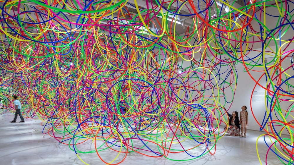 A large white gallery space is filled with brightly colored hula hoops, which are interconnected. Hoops skim the floor and hang from the ceiling. While there is a central mass of hoops, they also line some walls of the gallery. There are a few, tunnel-like pathways through the hoops. In one of these pathways a mother crouches down next to her young child, pointing up at the sculpture of hoops.