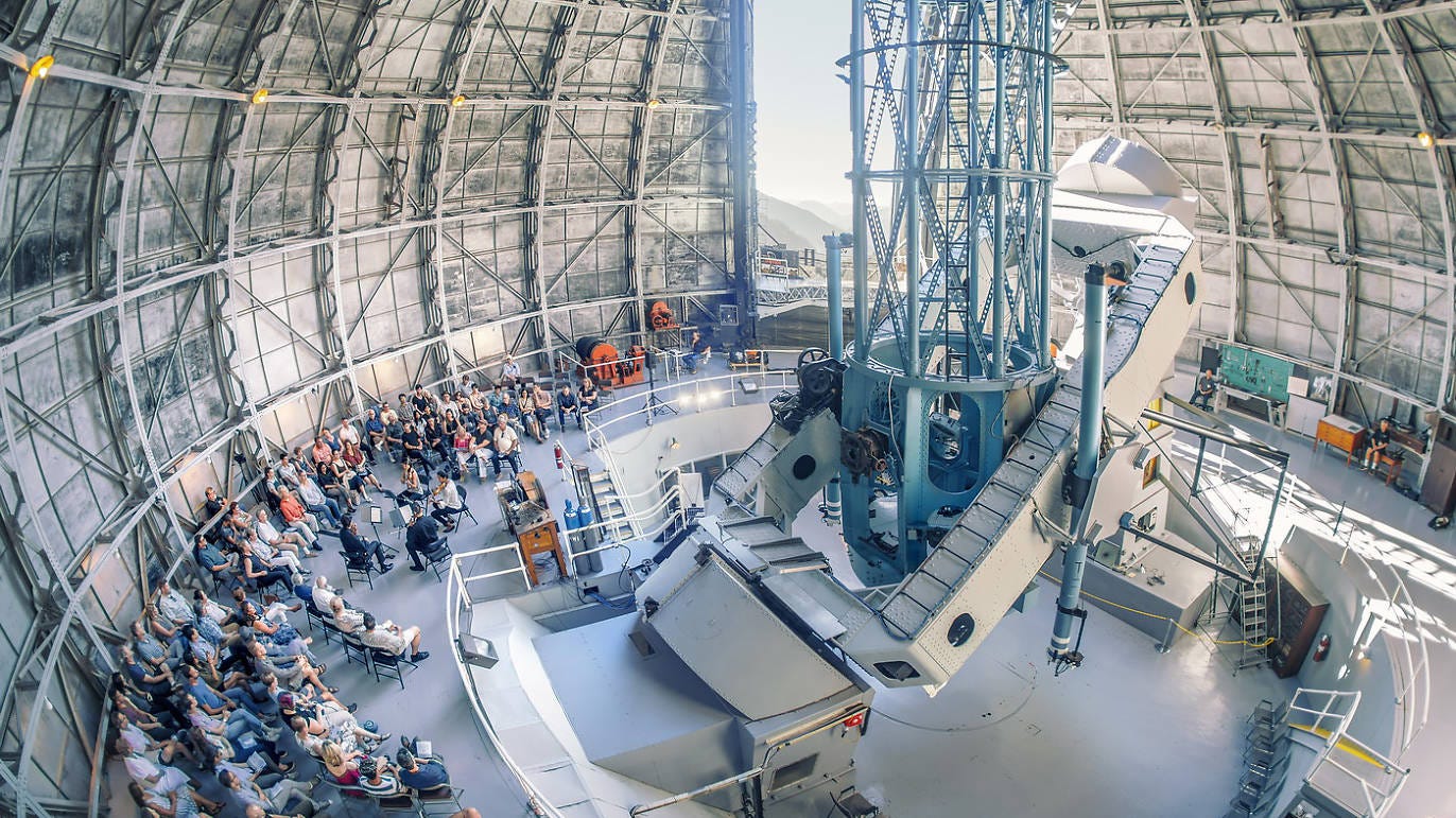 The interior of a massive telescope dome. Only a portion of the spherical walls are visible. The camera’s perspective is hovering above a group of people sitting and gathered around a presentation. The dome’s mechanical walls are partially open, revealing a slice of sunlight sky.