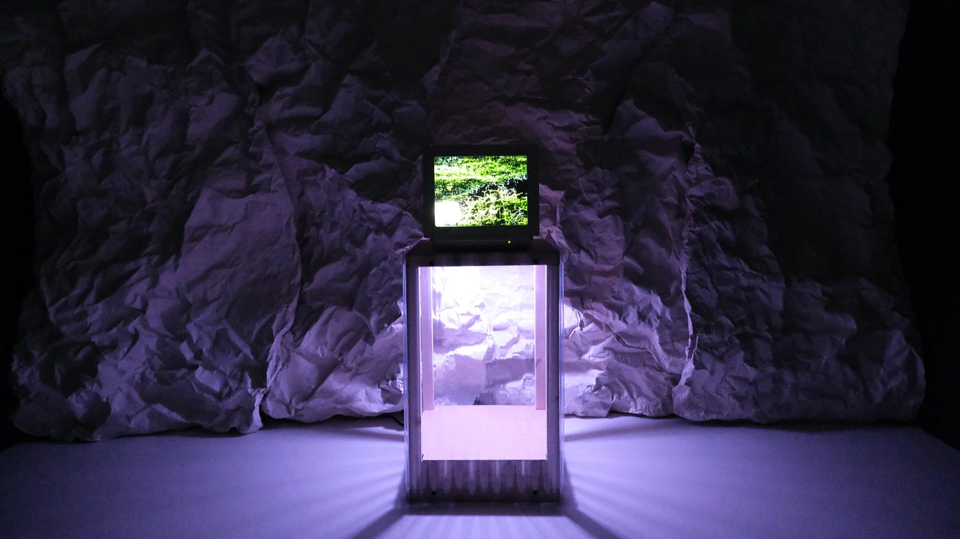 In the center of the image is a small tube television sitting atop a glass pedestal. There’s a verdant vista on its screen. The room surrounding it is dark, with an eerie, purple light emitting from the back of the TV. That light illuminates a back wall, which looks like a rocky face or the inside of a cave.