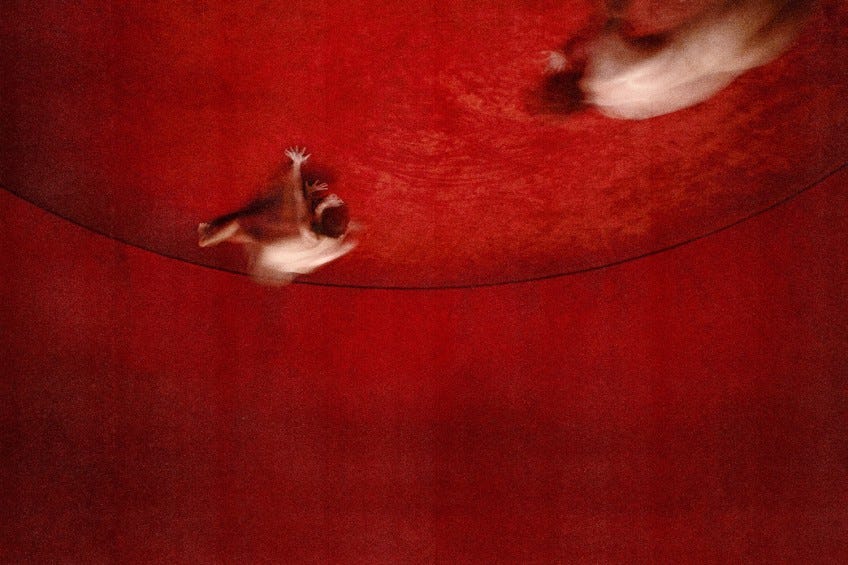A curved room completely covered in lush, red velvet. On the ceiling, two blurred, nude figures crawl towards each other.