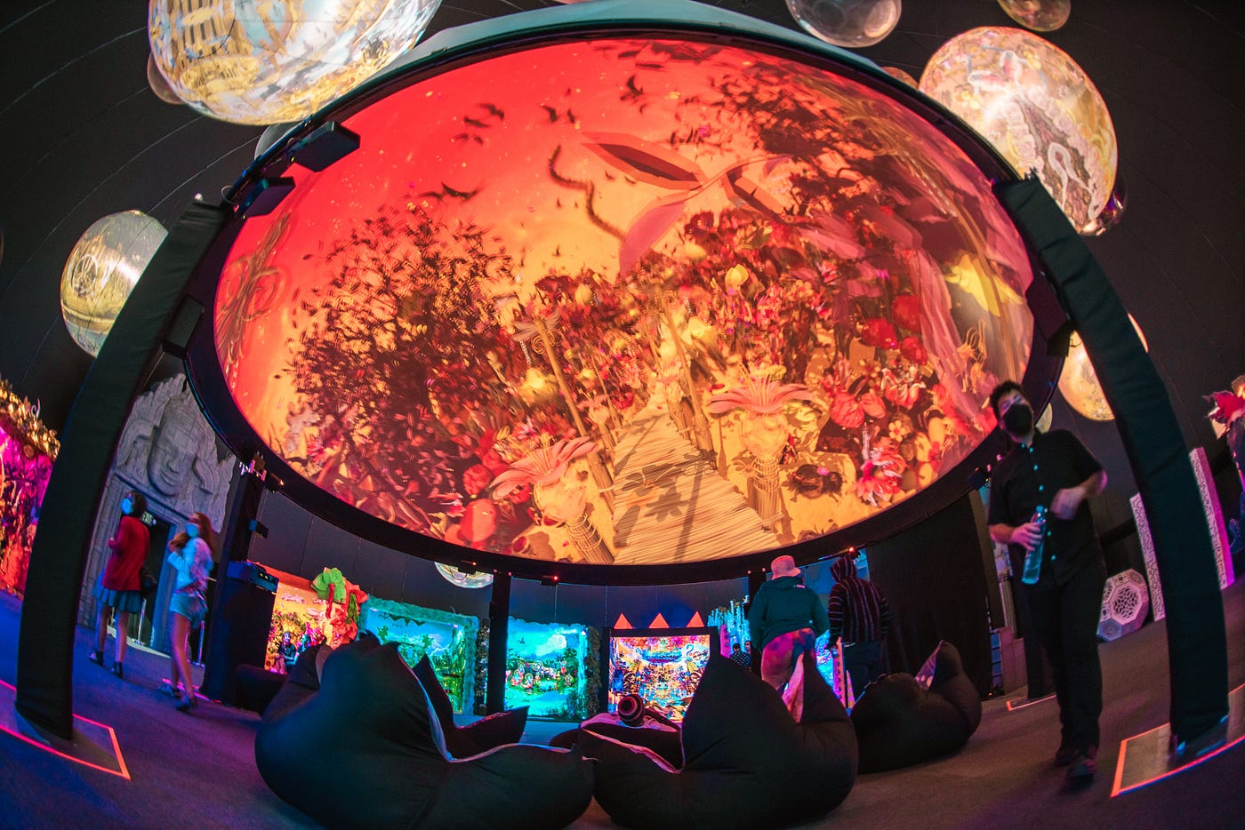 The image has a distorted view, curving the objects within. The central focus is the underside of a dome, which has digital art projected onto it. Bean bag-style chairs sit underneath. Other objects surround this main dome: grottos along the walls and inflated spheres, all are illuminated with bright colors and projections. Visitors stroll about.