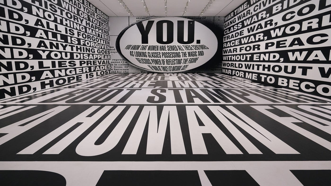 The view is from the center of a rear wall, looking at the surrounding walls and floor, which stretch far ahead. All of them are covered in massive, black-and-white text. The wall opposite has giant, slightly curved text encapsulated within an oval: “YOU. You know that women have served all these centuries as looking glasses possessing the magic and delicious power of reflecting the figure of man at twice its natural size.”