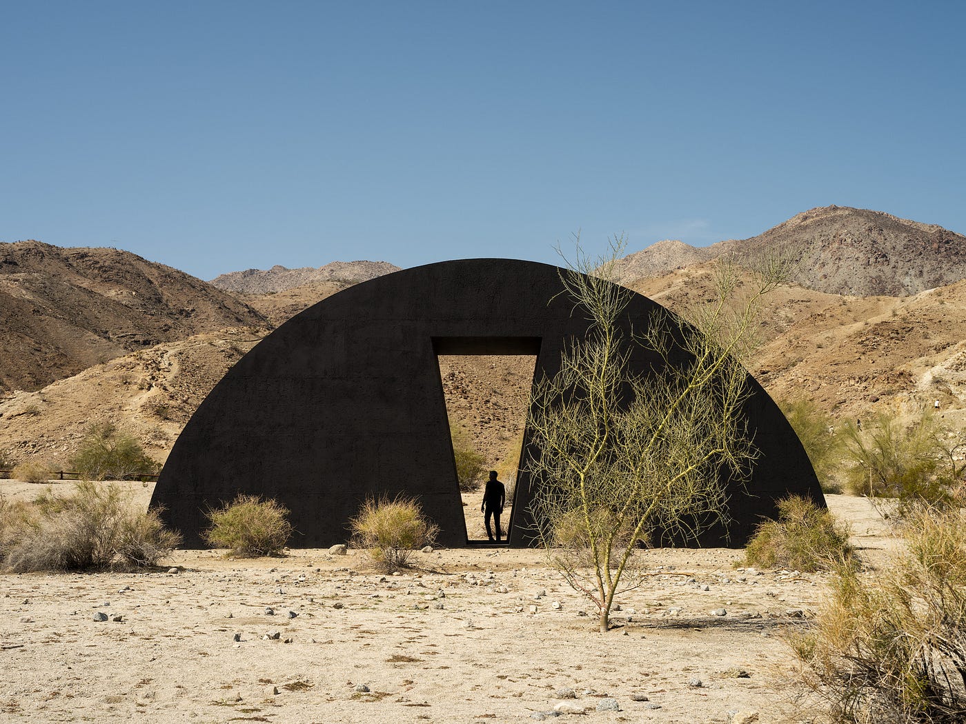 A giant, half-moon, black sculpture sits in the desert landscape. Behind it are sandy hillsides. At its center is an angular cutout, a doorway with the silhouette of a person standing inside.