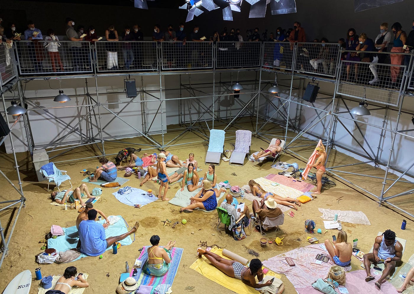 The setting is a fake beach. It’s inside a museum’s gallery. The “beach” is comprised of sand and performers are dressed in bathing suits, wetsuits, and casual summer garb. They lounge about on their towels or beach chairs, snacking, reading, and chatting. Surrounding them is metal scaffolding. Masked museum goers line the edges of the scaffolding, peering down on the scene.