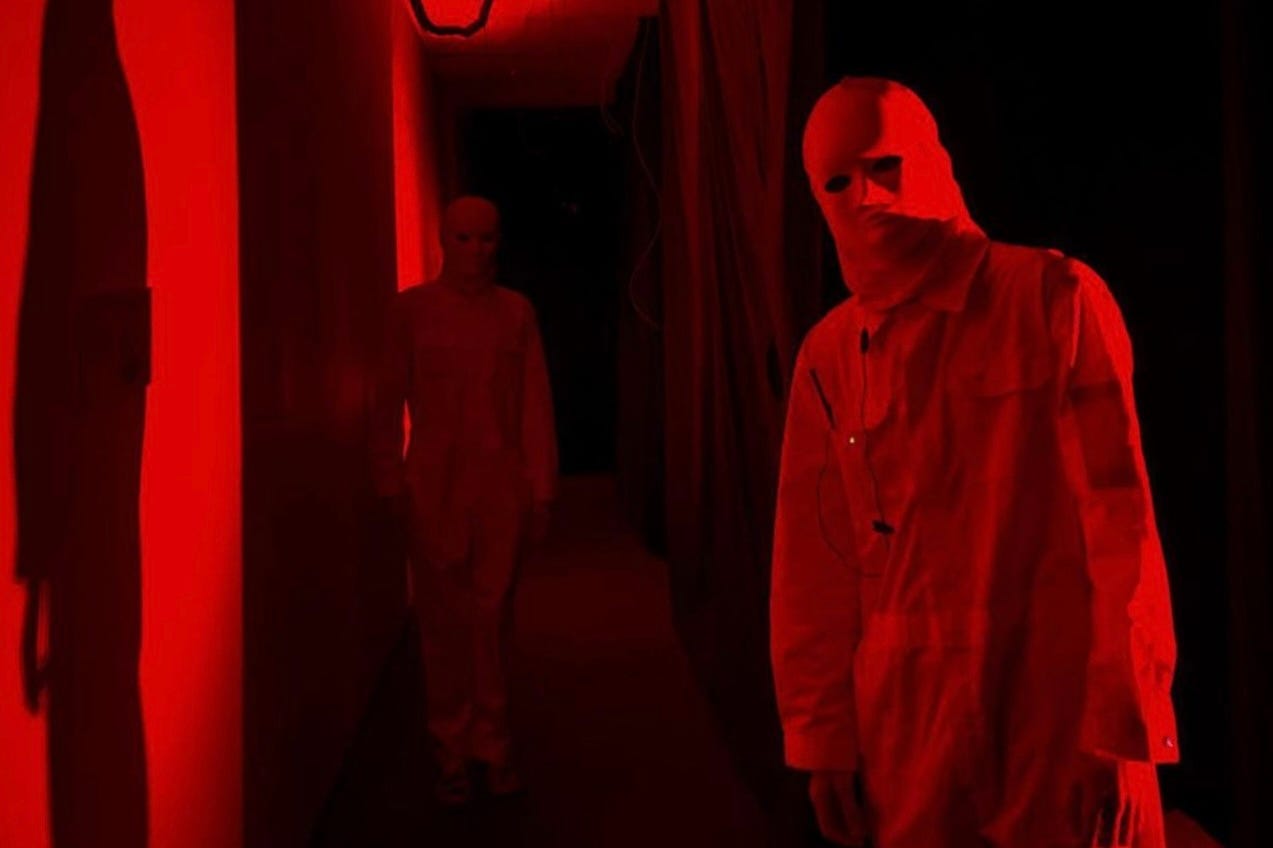 A darkened hallway illuminated by a dim, but saturated bath of red light. There are two figures, one you could reach out and touch, another further away. They are both dressed in white jumpsuits and wear white balaclavas.