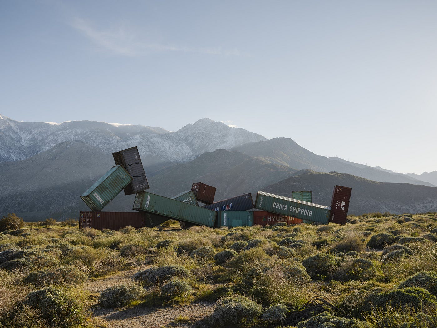 Snow-covered mountains in the distance. A group of massive shipping containers are arranged in the shape of a reclining figure. Its legs are crossed and the head is propped up by one bent arm.